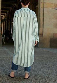 Lyseblå stribet skjortekjole med krave, lange ærmer og buet kant, parret med jeans med opsmøg og sorte slip-on sko.