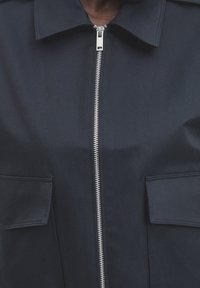 Granatowa wodoodporna kurtka z zapięciem na zamek, dużymi kieszeniami na piersi oraz usztywnionym kołnierzem z gładkiego materiału. Elegancki i nowoczesny design.