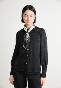 Blouse en satin noir avec un motif léopard texturé, manches longues, col en dentelle et boutons décoratifs en argent, associée à un foulard noir et blanc.