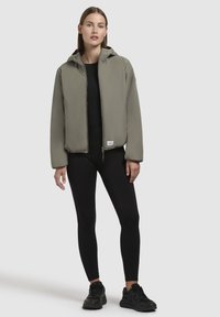 Light gray hooded jacket with a zip front, elastic cuffs, and a logo patch at the hem. Worn over a black fitted top and leggings.