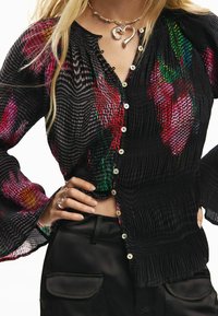 Schwarze, plisseeartige Bluse mit bunten Blumenmustern in Pink, Grün und Orange. Mit Knöpfen vorne und ausgestellten Ärmeln.