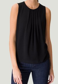 Schwarze ärmellose Bluse mit Falten am Ausschnitt, fließendem Stoff und entspannter Passform. Kombiniert mit dunkelblauen Jeans.