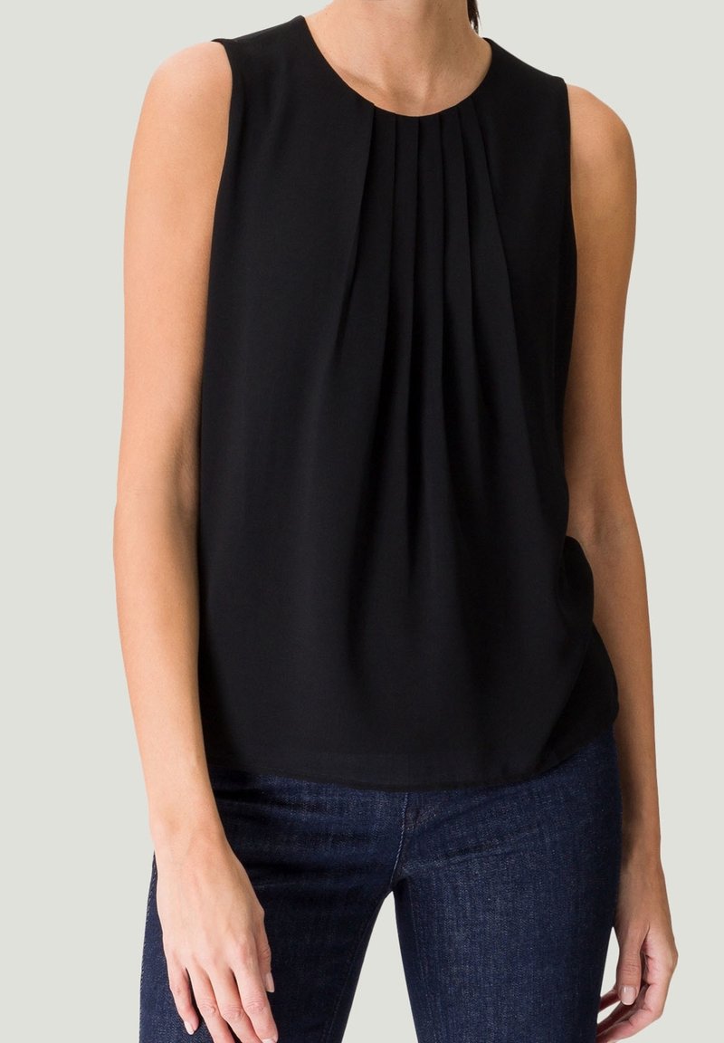 Schwarze ärmellose Bluse mit Falten am Ausschnitt, fließendem Stoff und entspannter Passform. Kombiniert mit dunkelblauen Jeans.