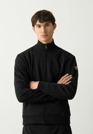 Jachetă neagră cu fermoar, guler înalt, mansete și tiv cu model în relief, textură netedă, având un logo rotund pe mâneca stângă.
