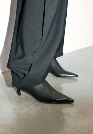 Czarne skórzane buty na kocim obcasie z zaostrzonym noskiem. Noszone z luźnymi czarnymi spodniami, prezentowane na betonowej powierzchni. Gładka faktura.