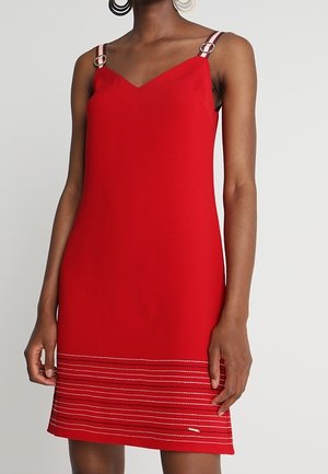 Frau trägt ein ärmelloses rotes Kleid mit dünnen Trägern und horizontalen weißen Nähten nahe dem Saum, stehend vor einem schlichten Hintergrund.