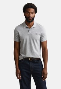 Man with beard wearing light grey short-sleeve polo shirt, dark jeans, and braided brown belt, standing against plain background.