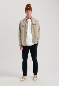 Camicia beige con bottoni, t-shirt bianca, pantaloni scuri, sneakers color sabbia; berretto in maglia marrone con un accento verde. Texture lisce, vestibilità casual.