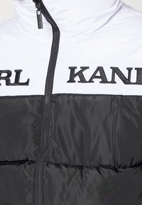 Padded jacket z czarną dolną częścią i białą górną częścią, z haftem 'KARL KANAI' w czerni oraz centralnym zamkiem błyskawicznym.