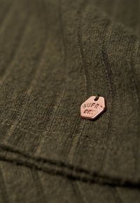 Tissu vert olive avec une texture côtelée, comportant une petite étiquette en métal rose avec l'inscription "SUPER DRY !" attachée au matériau.
