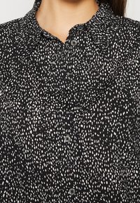 Chemise noire boutonnée avec motif irrégulier de points blancs et col large portée par une personne.