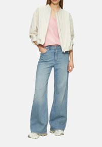 Hellbeige Bomberjacke, pinkes Zickzack-Muster-Oberteil und weit geschnittene blaue Jeans. Die Schuhe sind weiß mit beigen Akzenten. Lässiges Outfit.