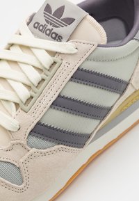 Ljust beige sneakers med grå ränder, överdel i tyg och mocka, gummisula, vita snören, adidas-logotyp på tungan och strukturerade accenter.