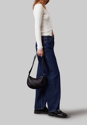 Musta, tikattu nahkainen olkalaukku, jossa on kaareva muoto ja säädettävä hihna, yhdistettynä vaaleaan ribattuun pitkähihaiseen puseroon ja leveälahkeisiin farkkuihin.