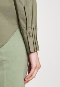 Camicia verde oliva con dettagli a pieghe sui polsini, dotata di due bottoni. Abbinata a una gonna liscia di un verde chiaro.