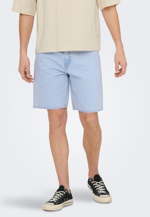 Mężczyzna ubrany w jasnoniebieskie dżinsowe szorty, beżową koszulę i czarne niskie trampki stojący na tle gładkiego jasnego tła.