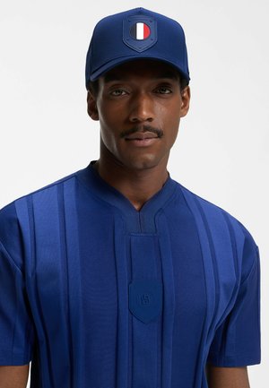 Mies, jolla on sininen lakki, jossa on punais-valko-sininen tunnus, ja vastaava sininen paita, jossa on pystysuoria raitoja ja rintataskun merkki.