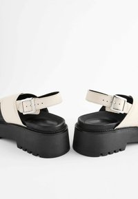 Beige platform-sandaalit, joissa on paksu musta pohja, nilkkaremmi, jossa on hopea-allas, ja tekstuuri yksityiskohtia remmeissä.