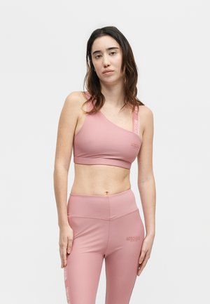 Woman wearing pink one-shoulder sports bra and matching high-waisted leggings with "MOSCHINO" logo on strap and thigh, standing against plain background.