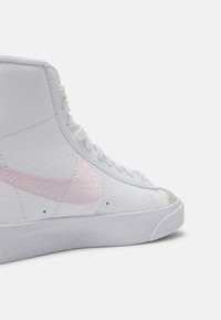 Zapatilla alta blanca con parte superior sintética texturizada, swoosh de Nike bordado en color rosa, cordones blancos y puntera en color gris claro.