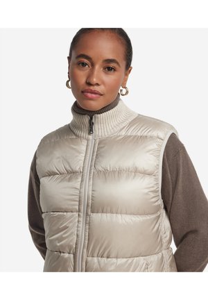 Kobieta w złotych kolczykach-kołach, ubrana w beżową pikowaną kamizelkę na brązowym golfie, patrząca na aparat z neutralnym wyrazem twarzy.