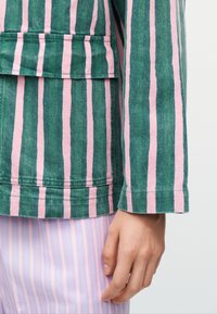 Mână odihnindu-se lângă o jachetă verde cu dungi verticale roz și pantaloni lila cu dungi verticale roz deschis.