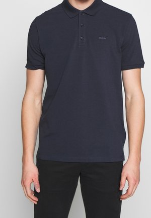 Man wearing a plain dark navy polo shirt with three buttons and black pants, standing against a plain light background.