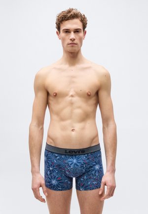 Young man with curly hair wearing blue floral patterned Levi's boxer briefs, standing against a plain light background.