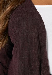 Burgundy kardigan med ribbad textur och öppen framsida, lager på lager över en vit topp. Tyget verkar mjukt och lätt.