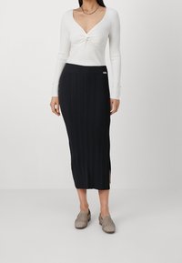 Biała bluzka z długim rękawem i skręconym przodem, w połączeniu z czarną, prążkowaną spódnicą maxi z bocznymi rozporkami. Modelka nosi tkane buty wsuwane.