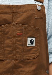 Καφέ παντελόνι εργασίας Carhartt με τσέπη και λευκή ετικέτα λογότυπου, μεταλλικό κλιπ, φορεμένο πάνω από ένα ανοιχτό μπλε πουκάμισο με υφή.