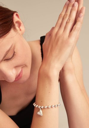 Pulsera de plata hecha de cuentas redondas, con un pequeño colgante en forma de corazón. El diseño es simple y pulido, descansando sobre una muñeca.