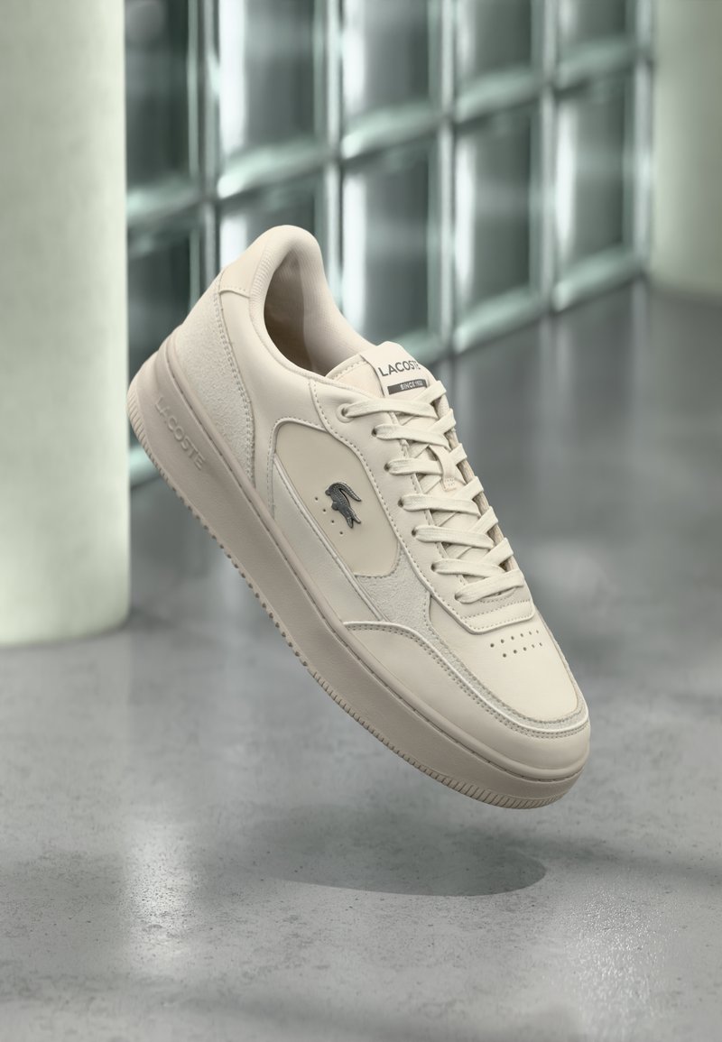 Baskets Lacoste blanc cassé avec lacets, logo en relief sur la semelle et emblème de crocodile sur le côté, présentées sur un fond moderne minimaliste.