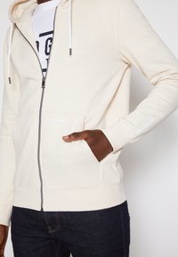 Personne portant un sweat à capuche crème zippé, main dans la poche avant et jean foncé, montrant les cordons de la capuche et une partie du t-shirt blanc imprimé.
