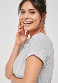 T-shirt gris à manches courtes avec un col rond, texture douce et poignets retroussés. Le mannequin a les cheveux raides et foncés sur un fond neutre.