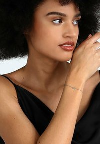 Silverarmband med delikat kedja och liten accent, buren på ett bart handled, med bäraren i en svart spaghetti-top.