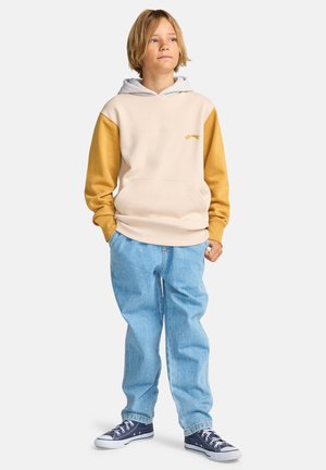 Beiger Hoodie mit gelben Ärmeln und grauer Kapuze, mit einer Fronttasche. Kombiniert mit hellblauen Jeans und dunkelblauen Sneakers.