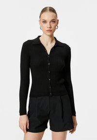 Schwarze gerippte Strickjacke mit Knopfverschluss und spitzem Kragen. Lange Ärmel und strukturierter Strickstoff, kombiniert mit schwarzen Shorts.