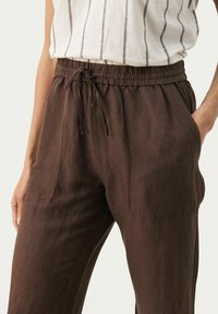 Bruna linbyxor med elastisk midja, dragsko och två sidfickor. Tyget har en mjuk, texturerad yta.