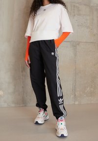 T-shirt bianca con logo nero, maniche lunghe arancioni, pantaloni da allenamento neri con strisce bianche e scarpe sportive multicolori con marchio.