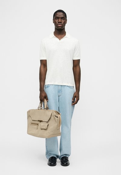 Άνδρας που στέκεται φορώντας λευκό πόλο με κοντά μανίκια, ανοιχτό μπλε τζιν, μαύρα παπούτσια, κρατώντας μια μεγάλη μπεζ τσάντα χειρός από τα χερούλια της.
