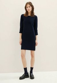 Kobieta stoi w granatowej sukience z długim rękawem, czarnych skarpetkach i masywnych czarnych loaferach na tle jasnym. 