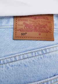 Jeans de denim azul claro con una etiqueta de cuero marrón que presenta gráficos en relieve, texto rojo y costuras doradas mostrando "501®" y otros detalles.