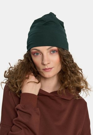 Kobieta z kręconymi włosami nosi ciemnozieloną czapkę i brązowy hoodie, trzymając pasmo włosów blisko twarzy na białym tle.