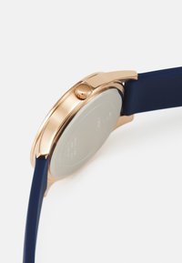 Reloj redondo con caja de acero inoxidable de color rosa dorado, correa de silicona azul y una corona texturizada. Diseño elegante con una parte posterior de acero inoxidable cepillado.