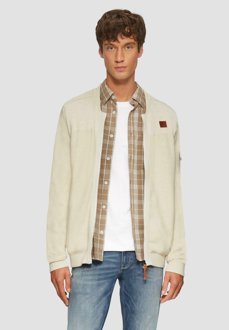 Hellbeige gestrickte Strickjacke mit gerippten Bündchen und Saum, unter der ein kariertes braunes Hemd und ein weißes T-Shirt getragen wird, kombiniert mit blauen Jeans.