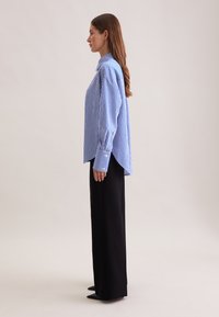 Camicia oversize a righe blu e bianche con colletto a punta e maniche lunghe, abbinata a pantaloni a gamba larga neri a vita alta e scarpe a punta.