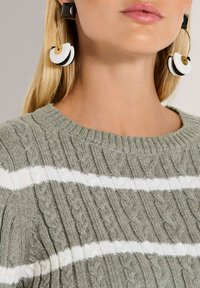 Strickpullover in olivgrün mit weißen Streifen; verfügt über ein strukturiertes Zopfmuster. Große schwarz-weiße Creolenohrringe mit goldenen Akzenten.