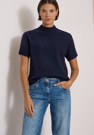 Femme portant un pull à manches courtes bleu marine foncé et un jean bleu clair taille haute, une main dans la poche et une expression neutre.