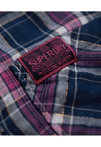 Rutiga skjortor i blått, lila och grått med en röd broderad "Superdry"-patch. Mjukt tyg med ett rutig mönster och synliga sömmar.